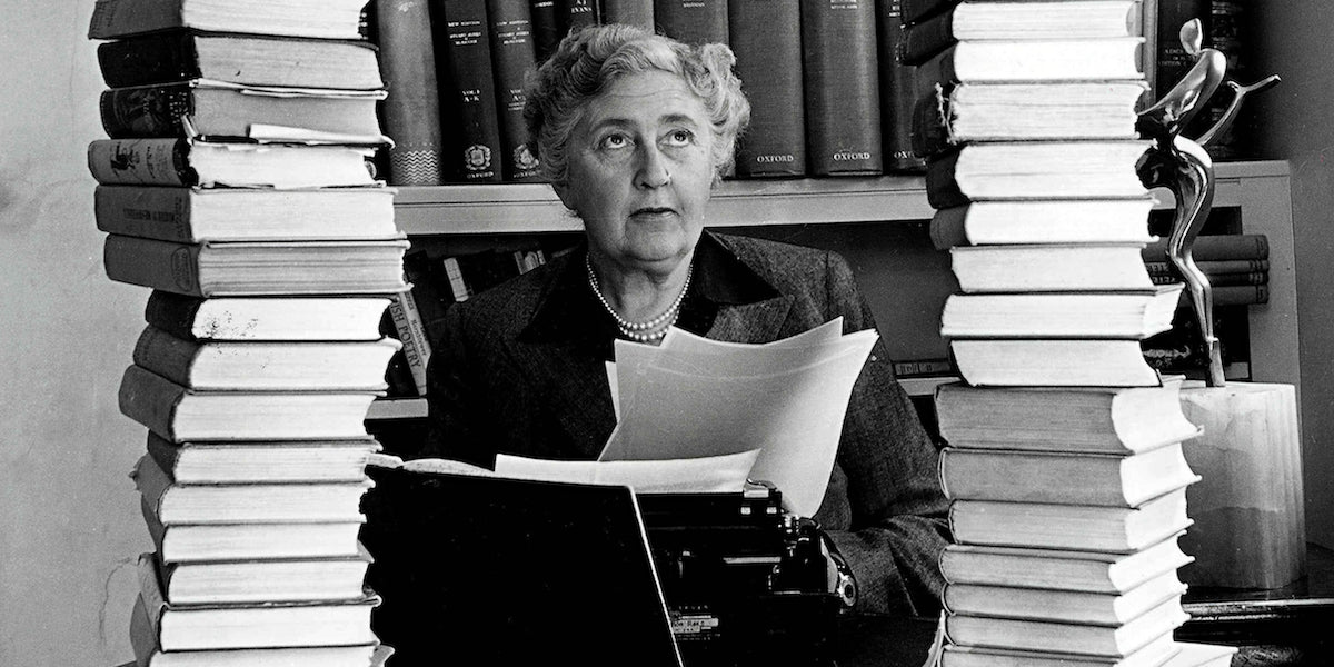 Black-and-white photograph of Agatha Christie seated at a desk, holding typed pages, framed by tall stacks of books in a study or library setting, with a thoughtful, focused expression.