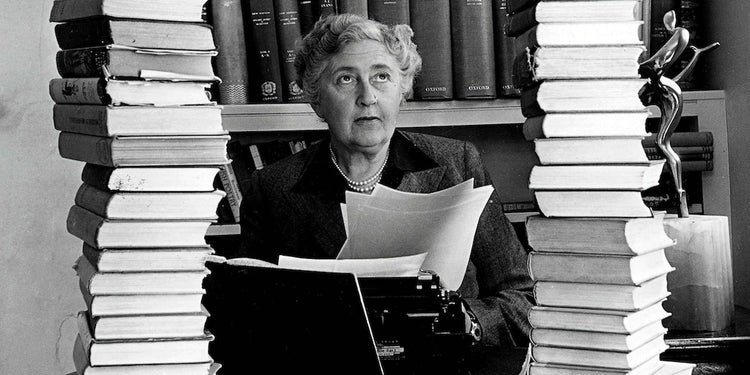 Black-and-white photograph of Agatha Christie seated at a desk, holding typed pages, framed by tall stacks of books in a study or library setting, with a thoughtful, focused expression.