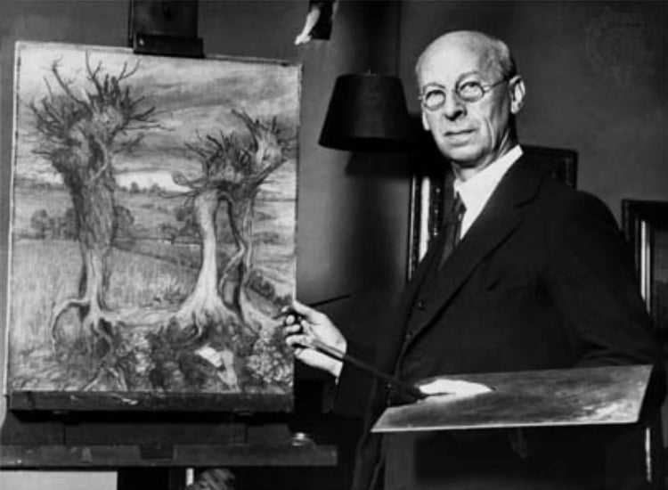 Black-and-white photograph of Arthur Rackham, illustrator standing beside an easel, holding a palette and brush, with a detailed woodland painting on display in a studio setting.