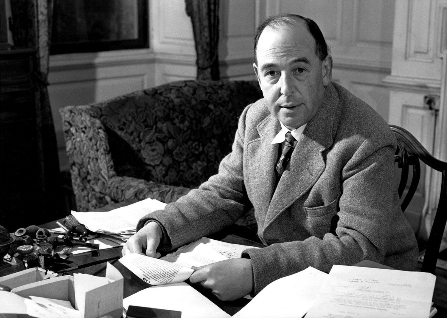 Black-and-white photograph of C. S. Lewis seated at a desk, reading and sorting manuscript pages, with papers and writing tools spread around him in an interior study setting.
