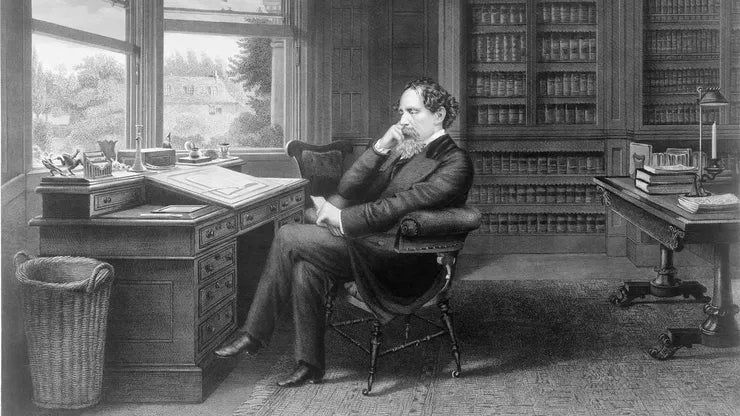 Black-and-white engraved illustration of Charles Dickens seated thoughtfully in an armchair at a writing desk, surrounded by bookshelves in a study or library.