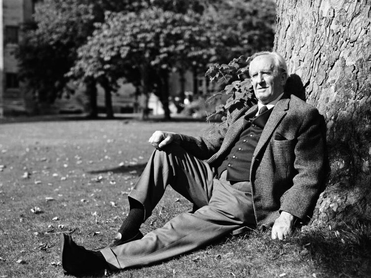 Black-and-white photograph of J. R. R. Tolkien in a tweed jacket seated on the grass, leaning against a tree in a park-like setting, relaxed and contemplative.